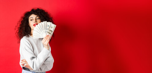 Wall Mural - Beautiful rich woman looking sensual aside, waving at herself with dollar bills fan, standing seductive on red background