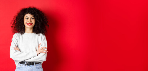 Wall Mural - Confident smiling woman with makeup and curly hairstyle, cross arms on chest and looking professional, red background
