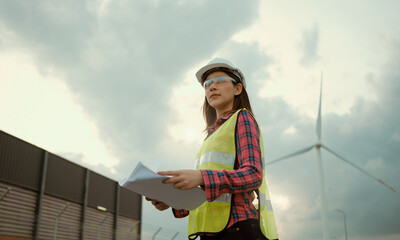 Asian woman engineer working and holding the report at wind turbine farm power generator station.