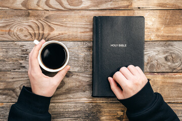 Bible and a cup of coffee in male hands on a wooden background, top view, concept of christianity and religion.