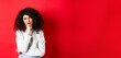 Grumpy young woman with curly hair, looking annoyed or bored at empty space, standing pensive and sad on red background