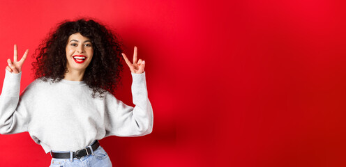 Wall Mural - Stylish young woman with curly hairstyle, smiling happy and showing peace signs, standing in casual sweatshirt on red background
