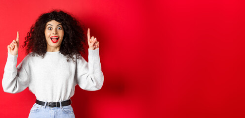 Canvas Print - Excited modern girl showing promo, pointing fingers up and smiling amazed, telling big news, standing on red background