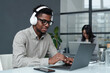 © AnnaStills - Young African American businessman in wireless headphones typing on laptop during his work at table in office