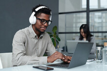 Young African American businessman in wireless headphones typing on laptop during his work at table in office