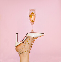 creative photo of a woman's foot in white shoes, with a glass of champagne on the shoe, on a pink ba
