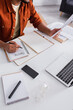 © LIGHTFIELD STUDIOS - Cropped view of copywriter holding paper and writing on notebook near devices and eyeglasses at home.