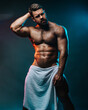 Big muscled man in white towel. Handsome hunk posing is studio. Naked male model with muscles and hairy body at dark background. Bodybuilder wearing only bath towel. Sexy athlete with six pack abs.