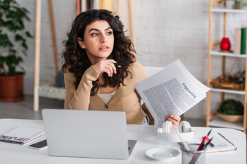 Brunette copywriter holding papers near gadgets and headphones in living room.