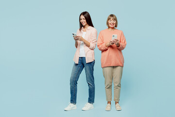 Wall Mural - Full body smiling happy cheerful elder parent mom with young adult daughter two women together wear casual clothes hold in hand mobile cell phone isolated on plain blue background. Family day concept.