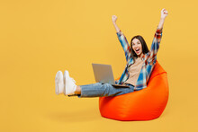 Full Body Young Happy Cool IT Woman Wear Blue Shirt Beige T-shirt Sit In Bag Chair Hold Use Work On Laptop Pc Computer Do Winner Gesture Raise Up Legs Finish Job Isolated On Plain Yellow Background
