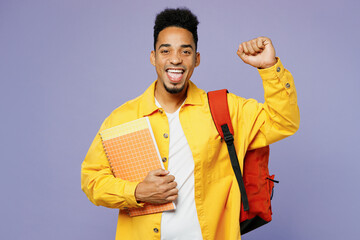 Young student man of African American ethnicity wear yellow shirt t-shirt backpack hold books do winner gesture isolated on plain pastel light purple background High school university college concept