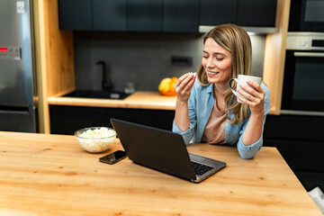 Wall Mural - A pretty blonde is standing in a modern kitchen checking emails on her laptop and online shopping, she's eating popcorn