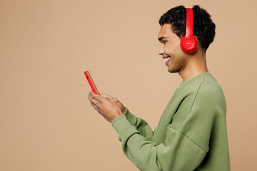 Wall Mural - Side view young man of African American ethnicity wear green sweatshirt headphones listen music use mobile cell phone isolated on plain pastel light beige background studio People lifestyle concept