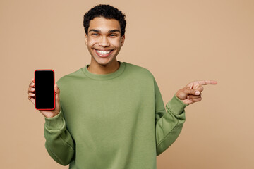 Wall Mural - Young man of African American ethnicity wearing green sweatshirt hold in hand use mobile cell phone with blank screen workspace area point finger aside isolated on plain pastel light beige background.