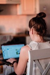 Young woman working remotely from home, sitting at table in kitchen using laptop, back view. Freelancer at home office, remote worker at workplace. Freelance and remote job concept