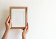 Woman's hands holding a wooden stylish blank frame against a white wall. Frame mock up, copy space for your text.	
