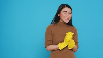 Wall Mural - Smiling asian maid clapping hands in front of camera after finishing to do house cleaning, standing in studio over blue background. Housekepper took great pride in her housekeeping duties