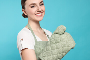 Wall Mural - Beautiful young woman in clean apron with pattern and oven glove on light blue background, closeup