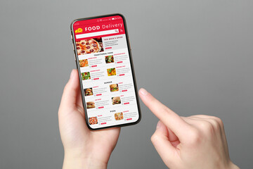 Food delivery service. Woman choosing dish from menu on site using smartphone on grey background, closeup