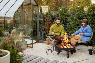 Young stylish couple grilling food and warming up while sitting together by the fire, spending autumn evening time at cozy atmosphere in garden