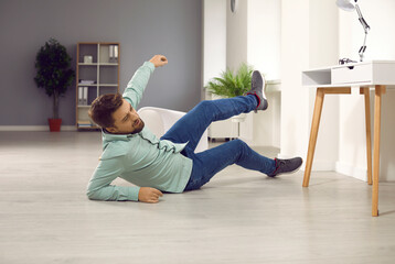 Full body photo of a young clumsy brunette man with unshaven bristles falling down to the floor and injuring his arm at home or in the office at his workplace and wearing casual clothes...