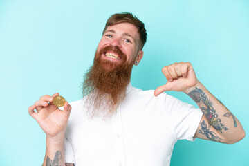 Wall Mural - Redhead man with long beard holding a Bitcoin isolated on blue background proud and self-satisfied