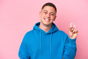 young brazilian man holding invisible braces isolated on pink background smiling a lot