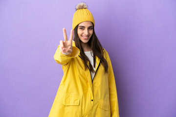 Poster - Young caucasian woman wearing a rainproof coat isolated on purple background smiling and showing victory sign