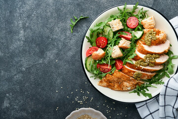 Wall Mural - Fresh salad with grilled chicken fillet, lettuce, arugula, tomatoes, cucumber and pesto sauce, sesame seeds with olive oil in white bowl on black slate background. Healthy lunch menu. Top view.