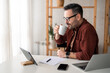 © Dorde - Freelance worker with glasses in casual clothing holding cup, drinking coffee in home office, sitting near window. Laptop computer, digital tablet device, smart phone, paper document and pen on table.