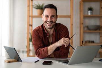 Smiling confident businessman looking at camera sitting at home office desk. Modern stylish corporate leader, successful manager or small business owner holding glasses posing for business portrait.