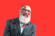 Senior Laughing Man With A Gray Beard In A Light Shirt In A Dark Casual Jacket With Lipstick Marks On Face Skin And Clothes. Bright Red Background