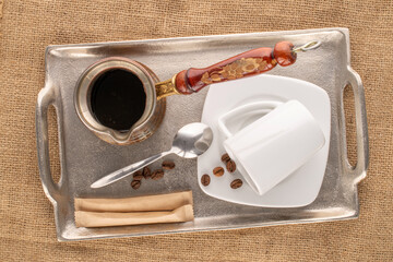 One cezve with coffee, white ceramic saucer with cup, metal spoon, several aromatic coffee beans and two packets of sugar with metal tray on jute cloth, macro, top view.