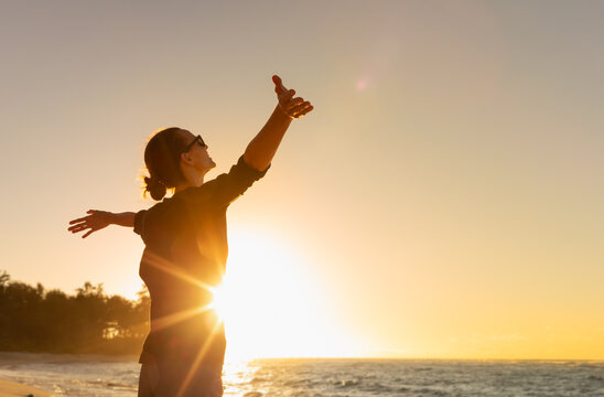 young woman facing ocean sunset rejoices, laughs, smiles looking up to the sky, enjoys life and summ