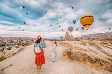 Wall Mural - Two girls friends capture the beauty of Cappadocia, Turkey, with their smartphones amid stunning rock formations and hot air balloons.