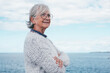 Portrait of pensive handsome senior woman standing by the sea looking away. Smiling elderly white-haired lady in outdoor excursion enjoying freedom, relax and retirement