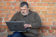 Nice portrait of bearded senior man working on a laptop and listening music while sitting against brick wall
