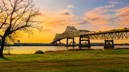 Horace Wilkinson Bridge over the Mississippi River green riverbank park with a barren willow tree at sunrise with golden glowing clouds on the blue sky in Baton Rouge, Louisiana, USA