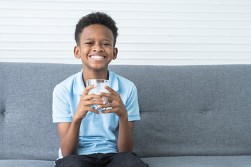 Portrait of adorable African Nigerian boy smiling with milk moustache, holding and drinking a glass of milk, sitting on sofa at home, looking at camera. Healthy food and drink for children concept