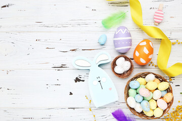 Wall Mural - Colorful eggs in baskets, clothespins, ribbon, feathers and rabbit on white wooden background