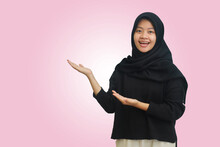 Portrait Of Confused Asian Hijab Woman In Black Outfit Standing Against Pink Background, Smiling And Looking At The Camera Pointing With Two Hands And Fingers To The Side