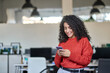 © insta_photos - Smiling young professional latin business woman, happy lady corporate leader holding cellular phone working standing in modern office using mobile apps cellphone technology device looking at cell.