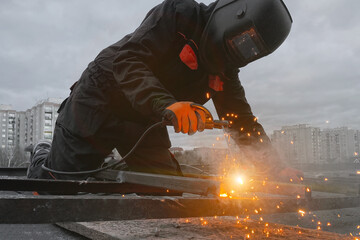 A welder in a suit and mask welds metal against the sky, welding work, a lot of sparks