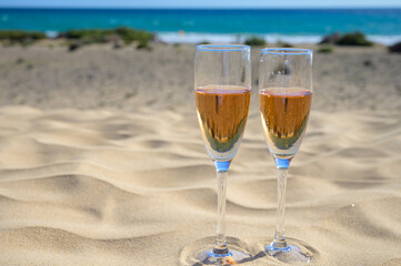 Two glasses of rose champagne or cava sparkling wine served on white sandy tropical beach with dunes and blue ocean, romantic vacation, winter sun on Fuerteventura, Canary, Spain