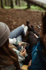 Wall Mural - Young european guy and lady tourists in jackets rest in autumn forest, enjoy cup of hot drink, free time in camping