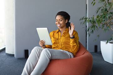 Wall Mural - Excited black businesswoman using digital tablet, sitting on pouf beanbag chair in office, having video call