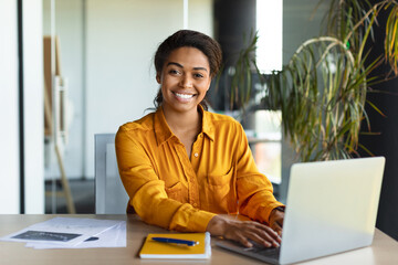 Wall Mural - Portrait of happy black businesswoman working on laptop and smiling at camera, sitting in office interior, copy space