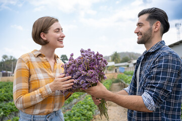 Man is holding purple flower bouquet to surprise his girlfriend at garden. Couple farmer harvesting strawberry in local farm with blue sky.