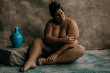 portrait of a plus size white woman sitting on the ground looking toward the sun holding her arm next to a blue vase